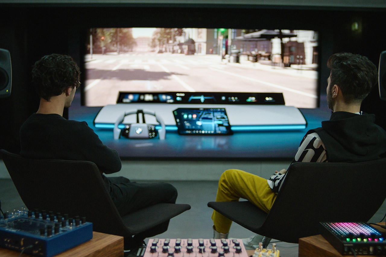 Two BMW Group sound designers in their studio, surrounded by tech, look at a virtual front screen with BMW Panoramic iDrive.