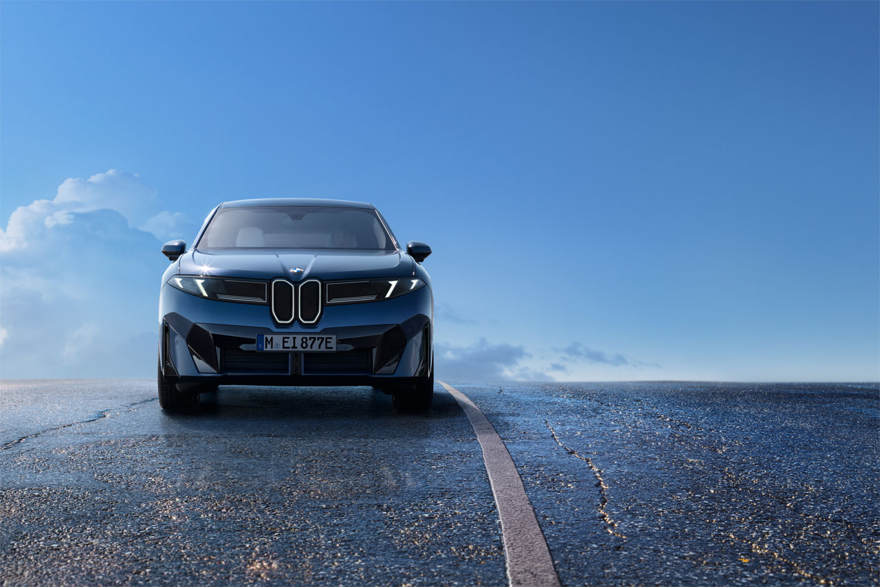 Front view of a dark-colored BMW iX3 on a road, with blue sky and clouds in the background.