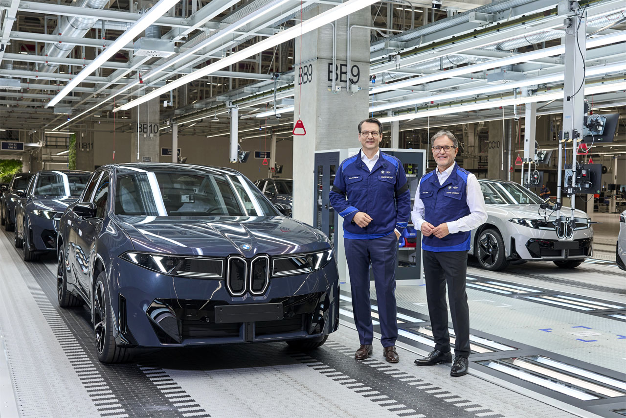Two men in blue work clothes are standing next to a black BMW sedan on the assembly line in a car factory.