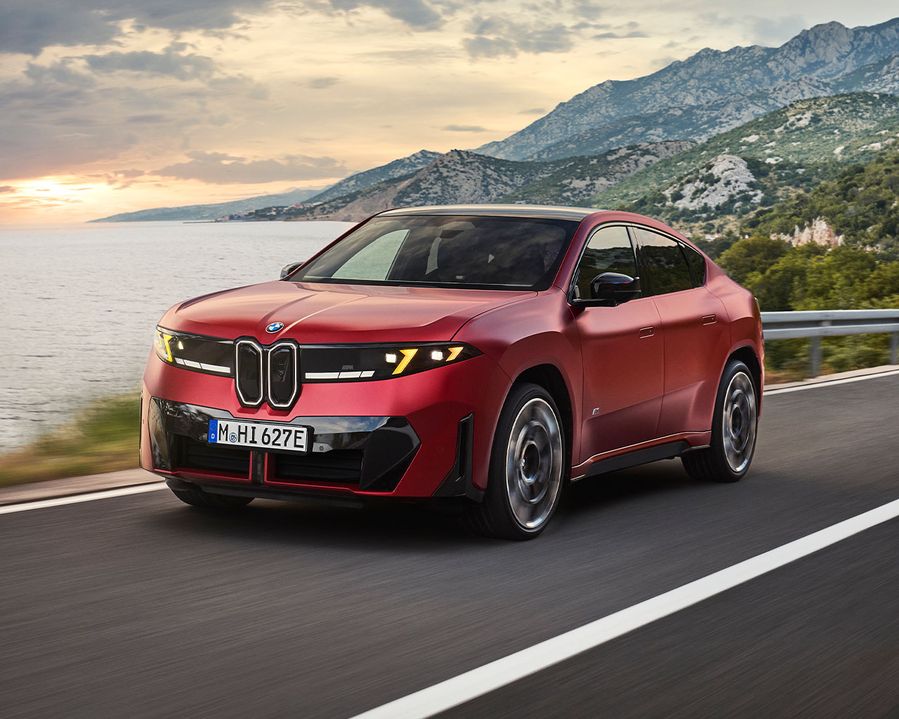 Schräg von vorn ist ein roter BMW iX4 M60 xDrive zu sehen, der auf einer Küstenstraße fährt. Im Hintergrund liegen das Meer und eine Bergkette im Abendlicht.