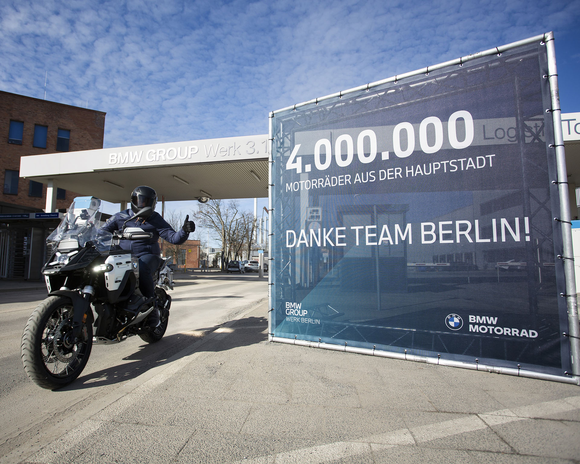 A motorcyclist leaves the premises of BMW Group Plant Berlin on a BMW R 1300 GS Adventure made at the facility. On the right, a large sign reads “4,000,000 motorcycles made in the capital – Thank you, Team Berlin!”