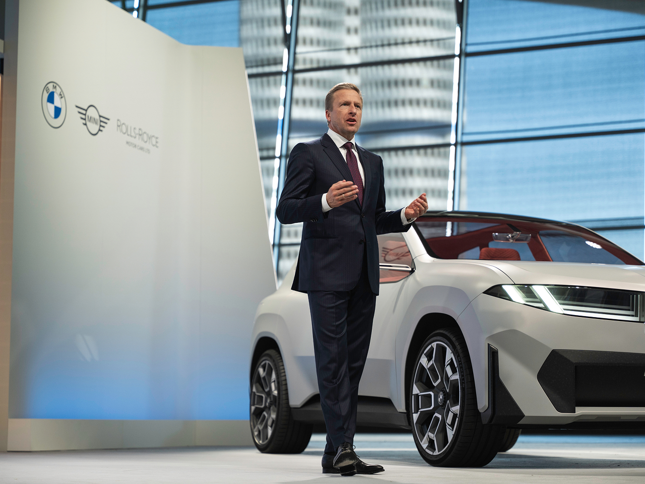 Oliver Zipse spricht auf einer Bühne neben einem silbernen BMW-Konzeptfahrzeug, im Hintergrund das BMW-Vierzylinderhochhaus.