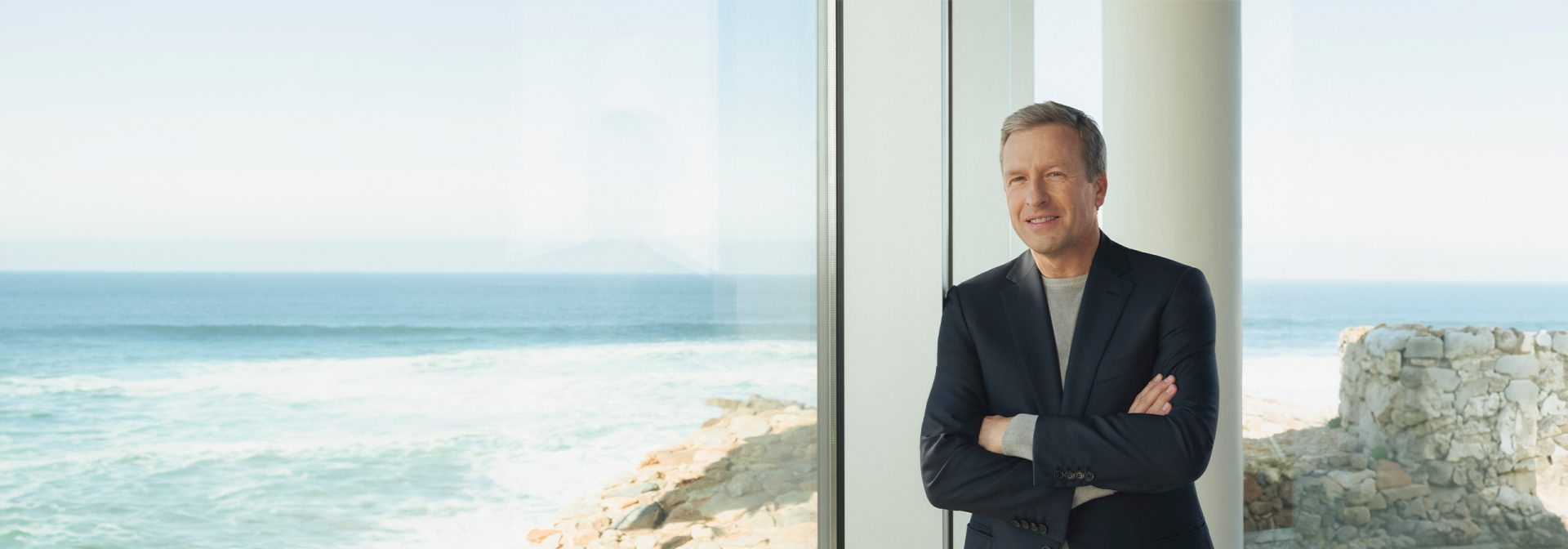 Oliver Zipse, der CEO der BMW AG, mit verschränkten Armen an einer Fensterfront stehend. Das Meer ist im Hintergrund.
