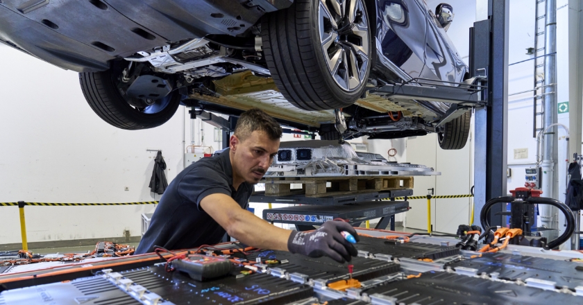 Ein Mann arbeitet in einer Werkstatt an einer technischen Platte. Über ihm hängt ein Auto.