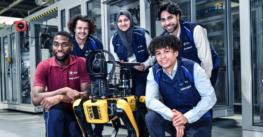 Fünf Menschen stehen in einer Werkshalle um einen Roboterhund herum und lächeln in die Kamera.
