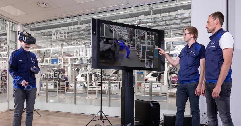 Ein Mann trägt eine VR-Brille, während die anderen beiden auf einen Monitor schauen.