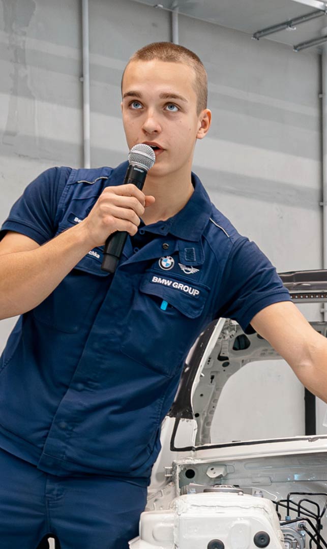 Person in BMW Arbeitskleidung erkl&auml;rt technische Komponenten im Motorraum eines freigestellten Fahrzeugs und spricht in ein Mikrofon.