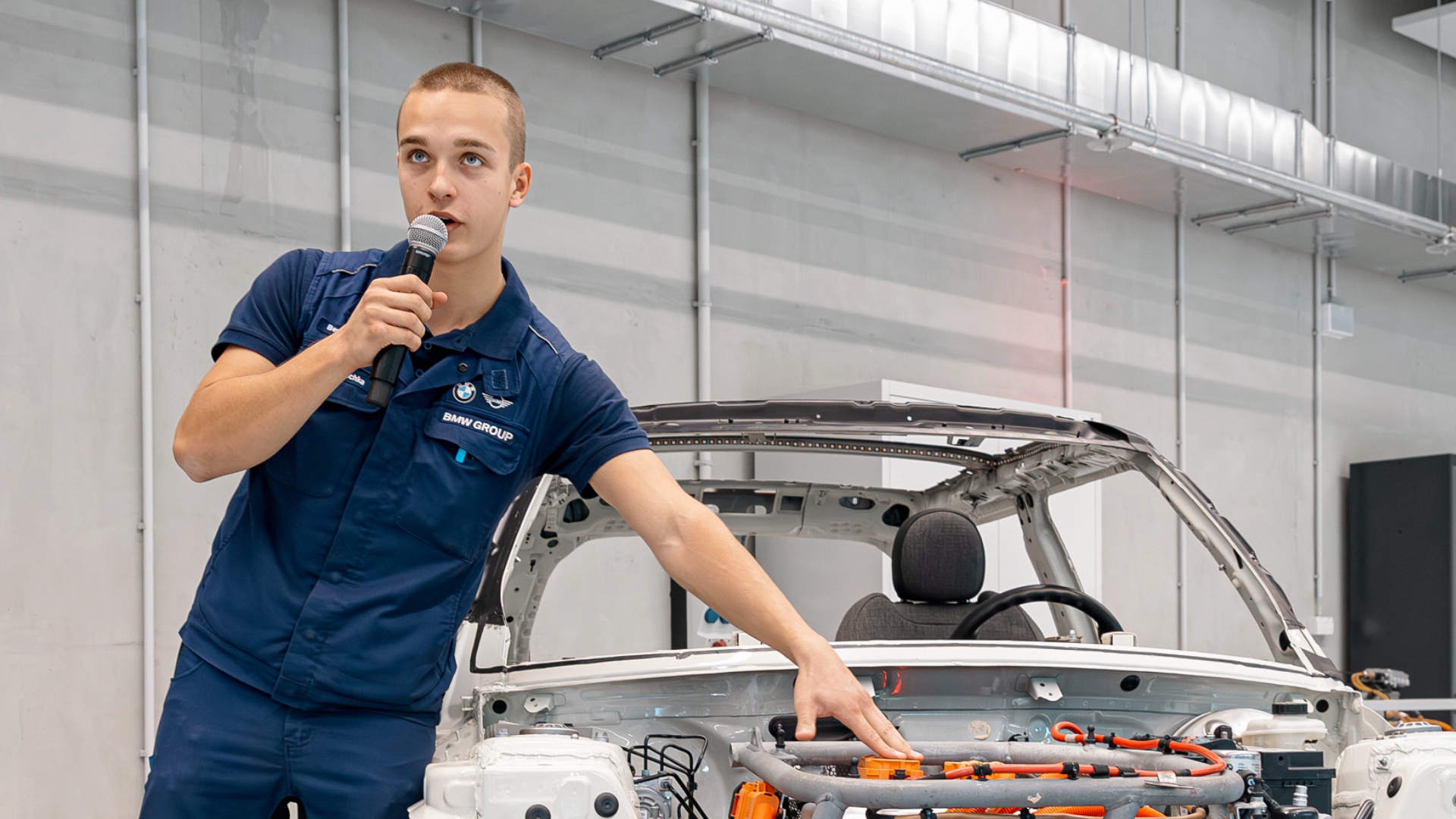 Person in BMW Arbeitskleidung erkl&auml;rt technische Komponenten im Motorraum eines freigestellten Fahrzeugs und spricht in ein Mikrofon.