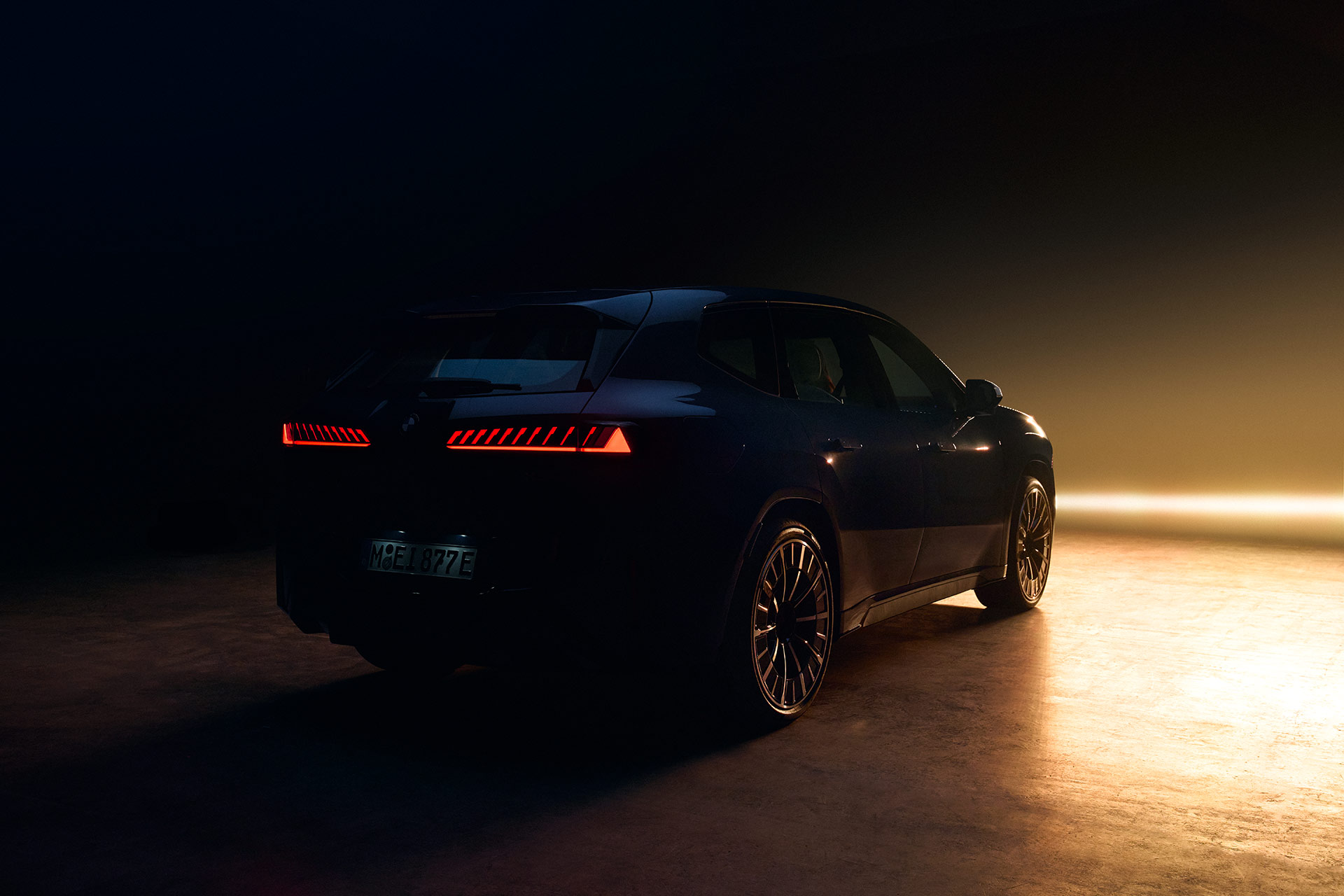 R&uuml;ckseite eines BMW Fahrzeugs in dunkler Umgebung, mit roten LED‑R&uuml;ckleuchten und warmem Licht im Hintergrund.