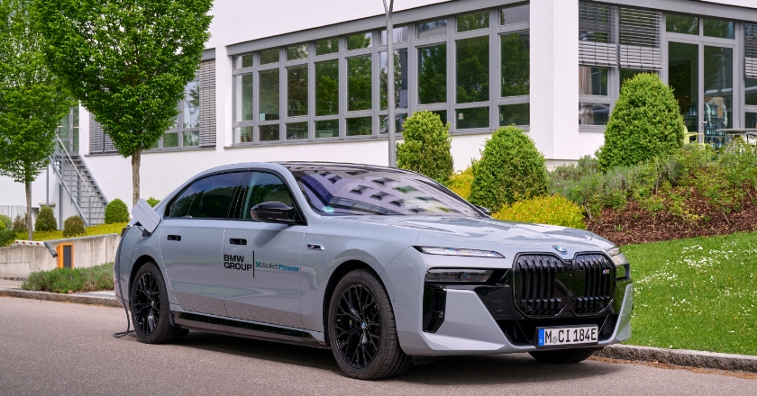 Elektrische BMW Limousine mit Solid‑Power‑Markierung beim Laden an einer Außenstation vor einem Bürogebäude.