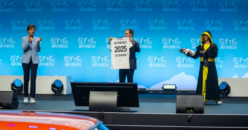 Drei Personen auf einer Bühne vor One‑Young‑World‑Hintergrund, eine Person hält ein Shirt mit der Aufschrift „One Young World 2025 Start Movement“.