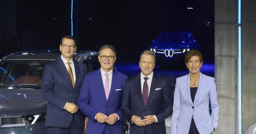 Vier Personen stehen zwischen mehreren BMW iX3 Fahrzeugen in einer Halle mit blau beleuchteter Hintergrundszene.