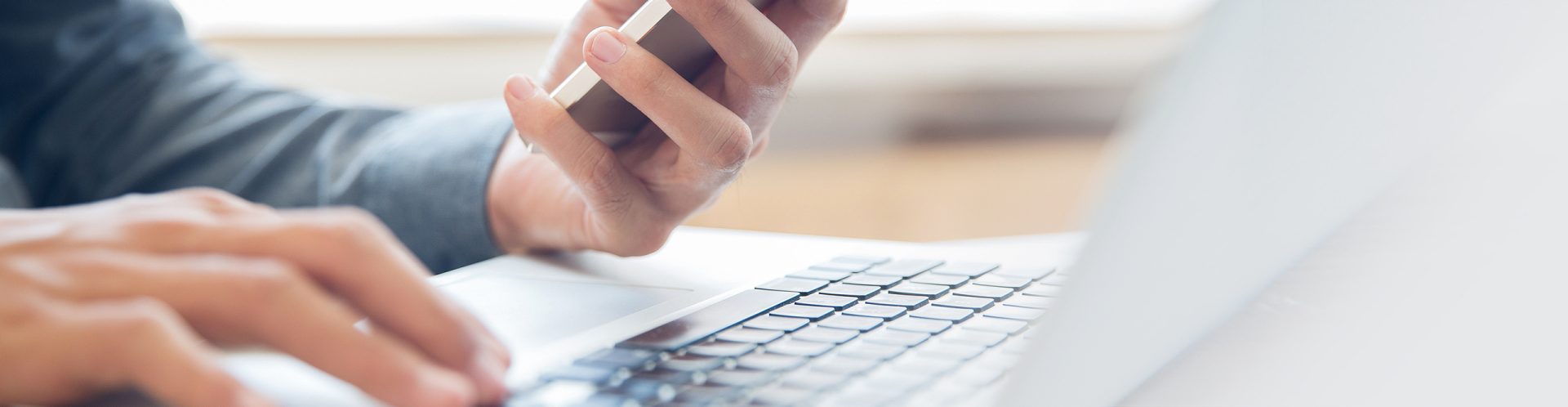 Eine Person hält ein Handy in der Hand und tippt etwas in den LapTop ein.