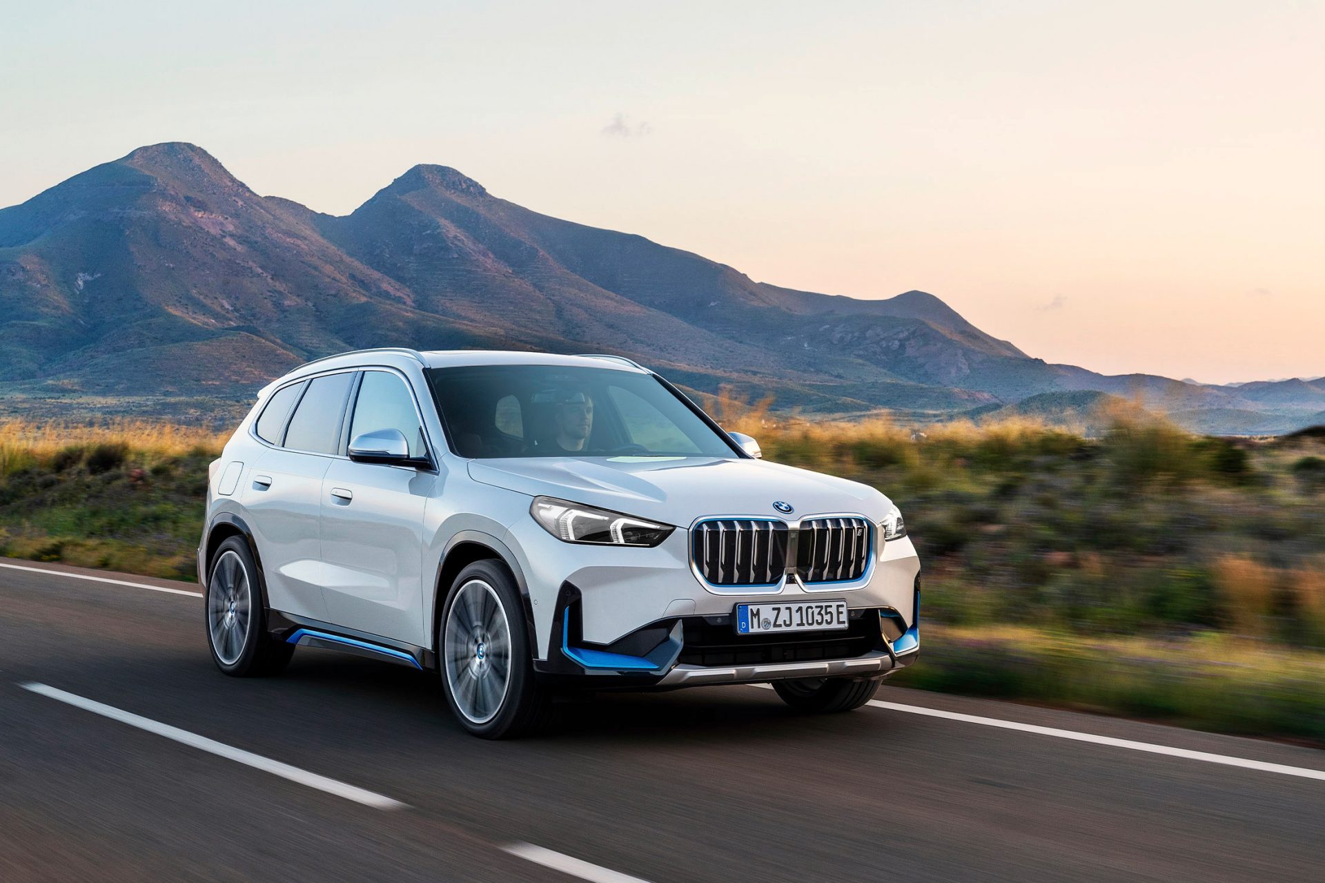 Weißer BMW iX1 fährt auf einer Landstraße bei Sonnenuntergang.