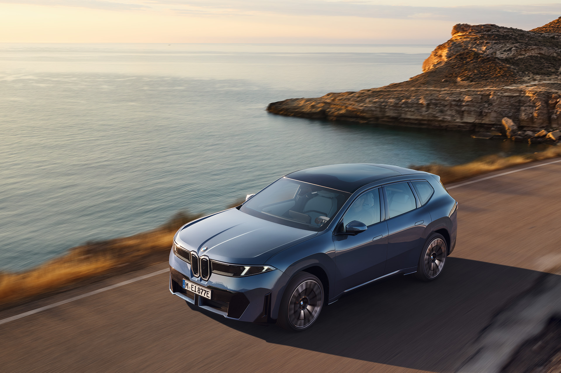 Ein blauer BMW iX3 fährt auf einer Landstraße am Meer entlang.