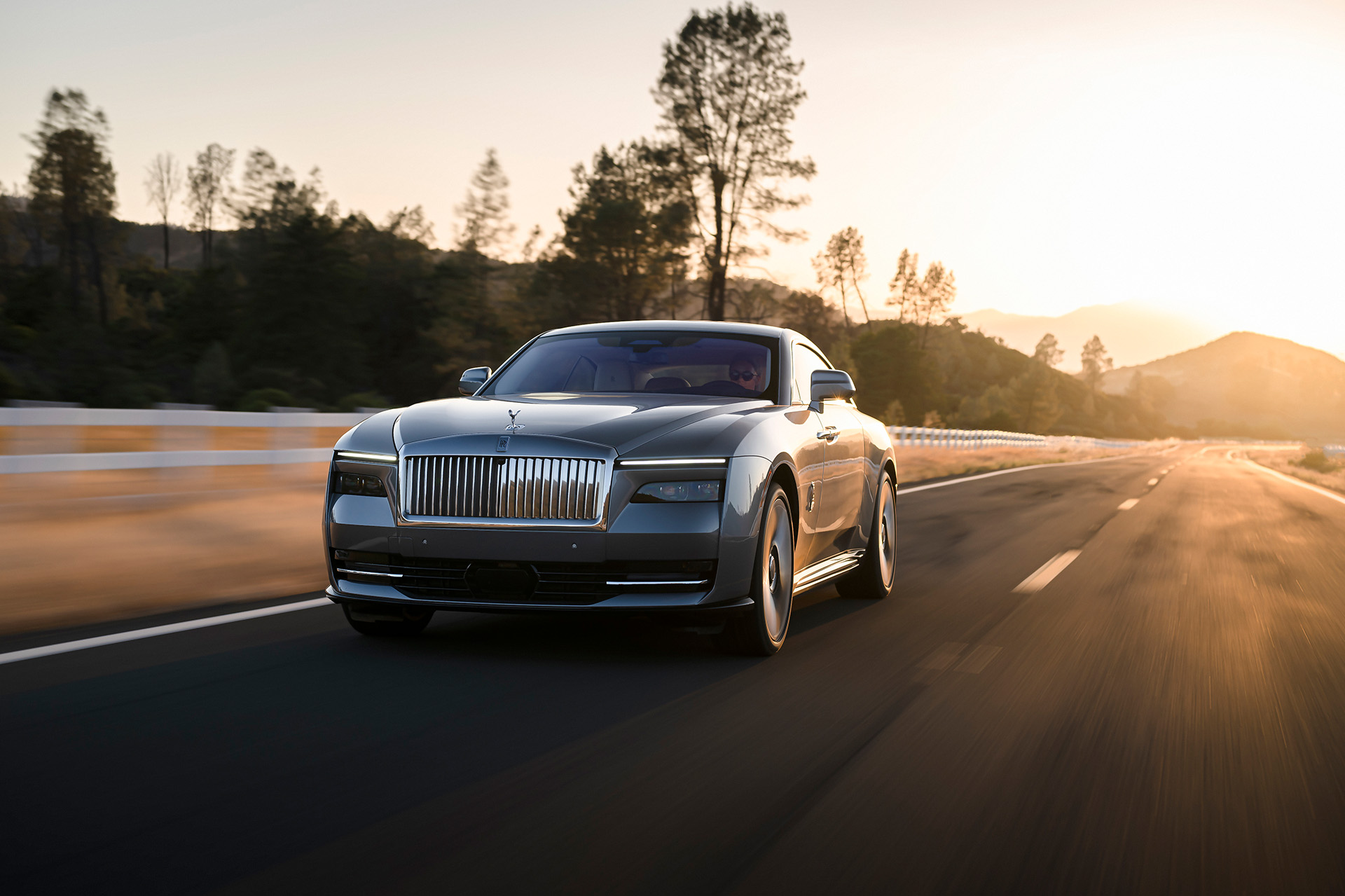 Ein grauer Rolls-Royce Spectre fährt bei Sonnenuntergang auf einer leeren Landstraße.