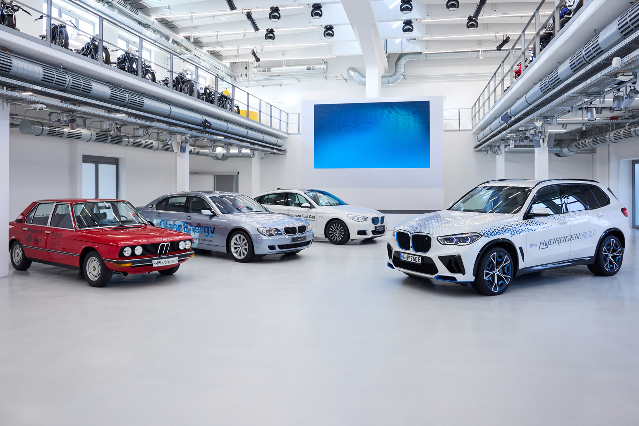 Vier BMW Wasserstofffahrzeuge von alt bis neu stehen in einer hellen Halle.