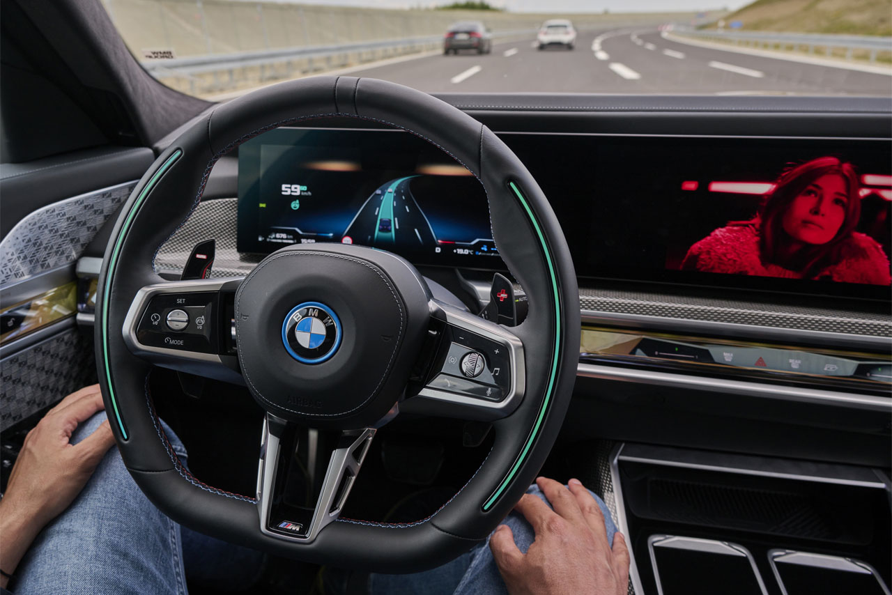 BMW-Lenkrad mit digitalen Anzeigen, Fahrer sitzt, Blick auf Autobahn mit mehreren Fahrzeugen.
