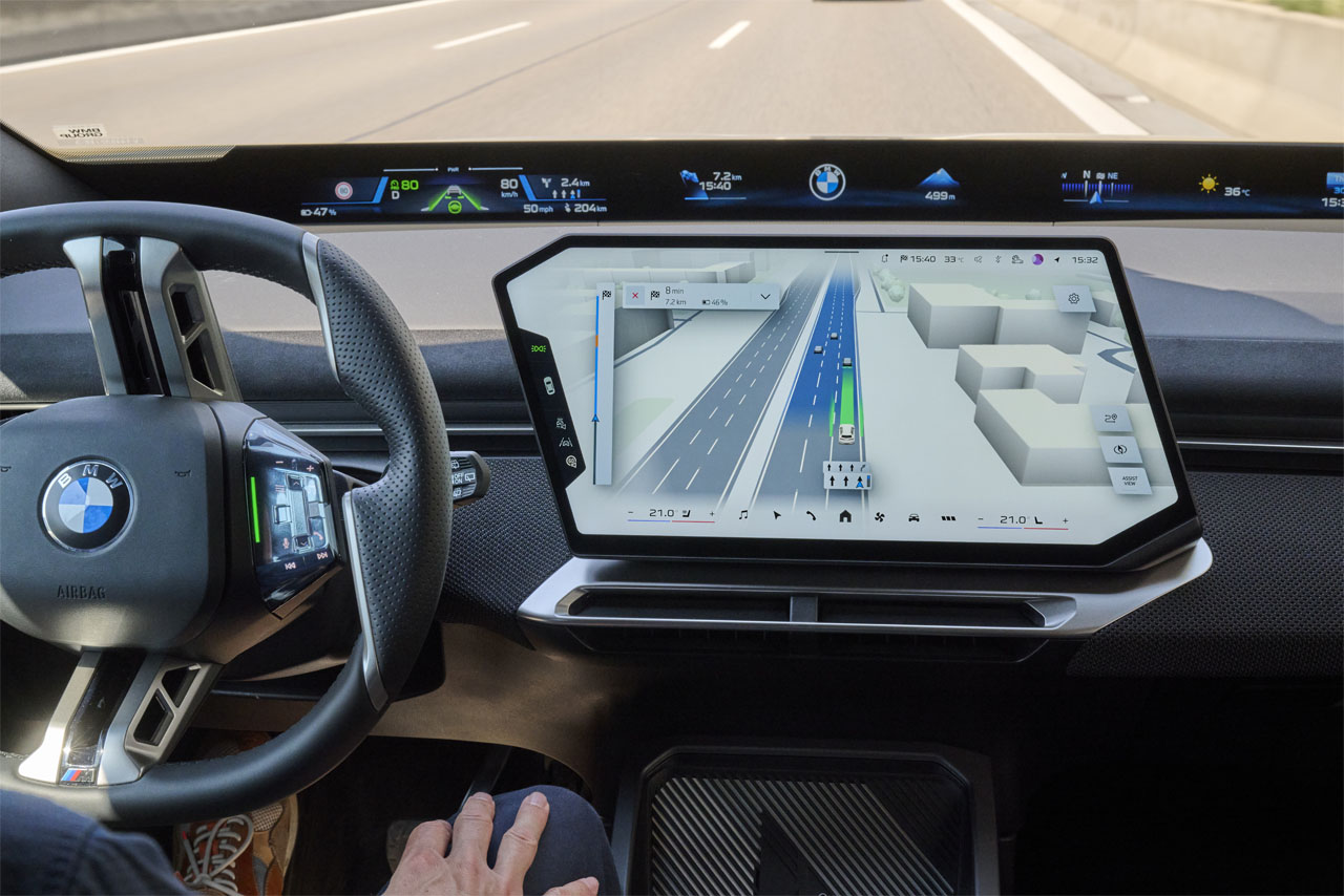 Blick ins Cockpit eines BMW iX3 auf der Autobahn: Das Zentraldisplay zeigt Route und Fahrerassistenz, die Hände des Fahrers ruhen im Schoß.