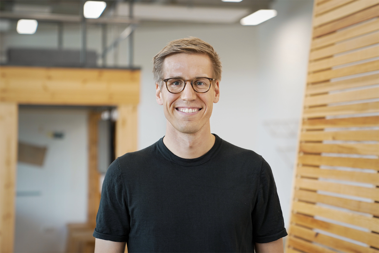 Mann mit kurzem, blonden Haar trägt ein schwarzes T-Shirt. Er steht in einem Raum mit Holzregalen und einem modernen, minimalistischen Design.