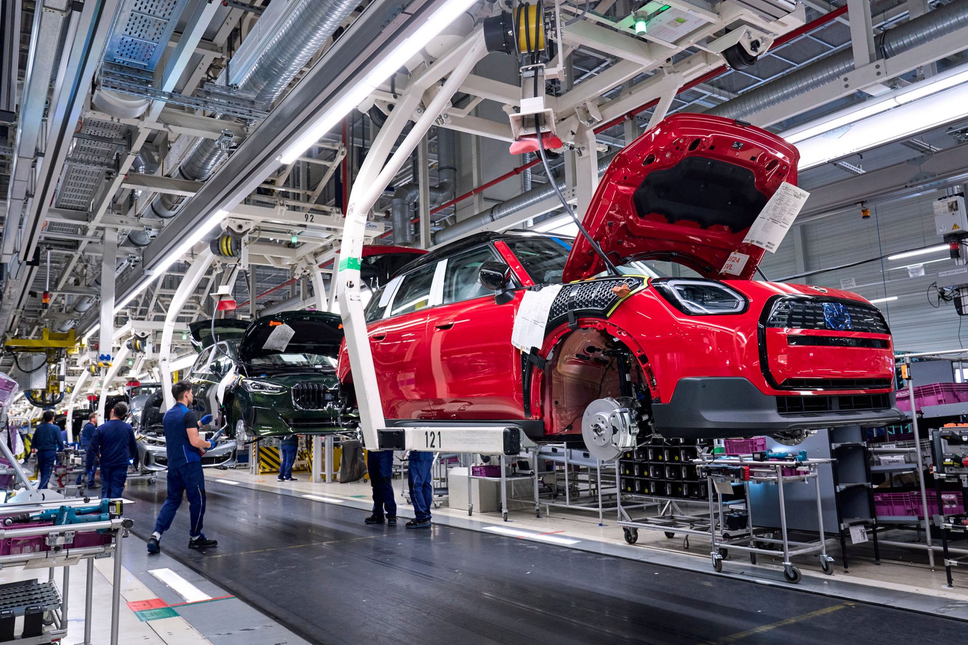 Hochautomatisierte Montagelinie im BMW Group Werk Leipzig: Ein roter MINI Countryman Electric und BMW Fahrzeuge hängen in der Fertigungseinrichtung, während Mitarbeitende an Karosserie und Komponenten arbeiten.