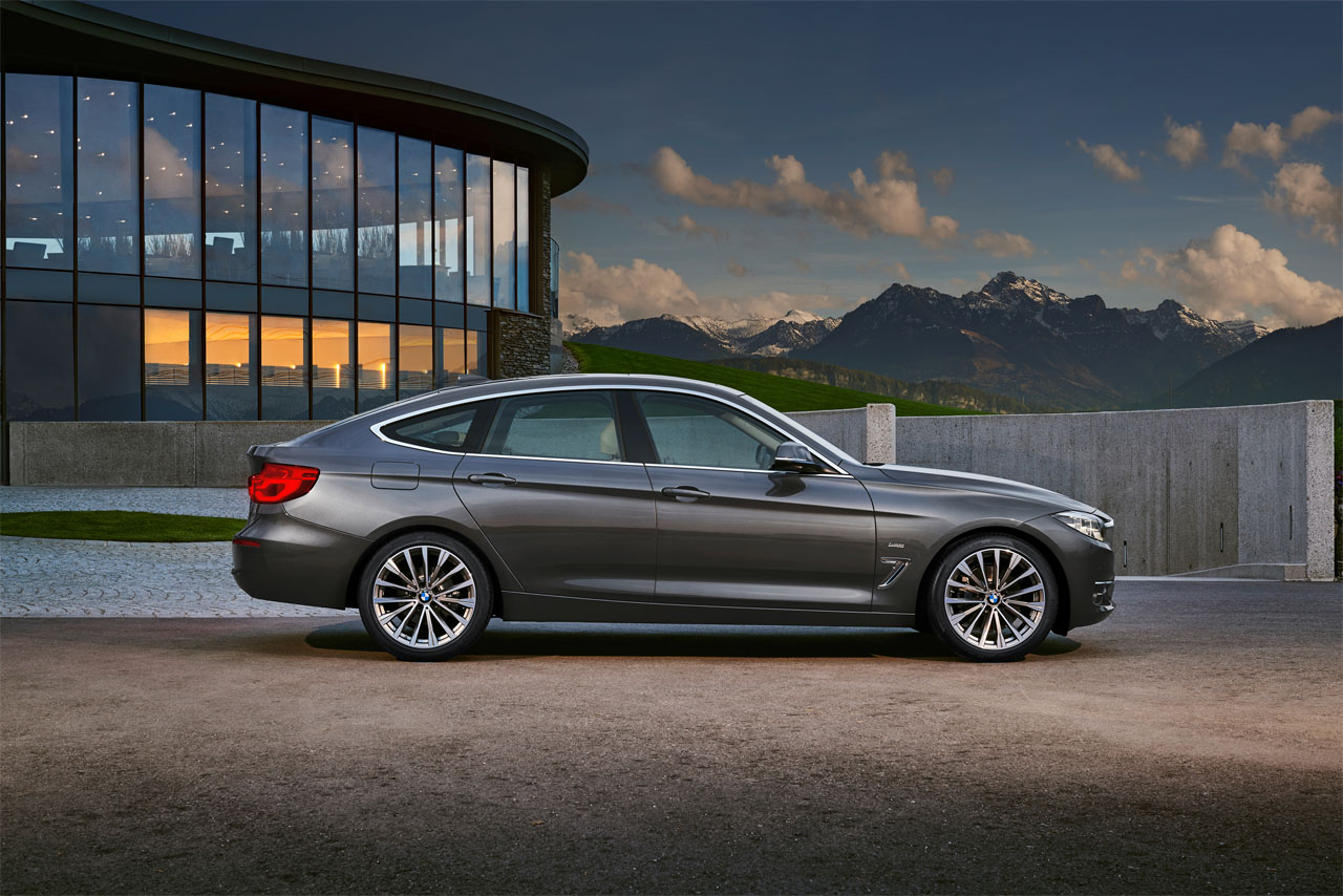 Ansicht auf die Beifahrerseite eines dunkelgrauen BMW F34 Gran Turismo vor moderner Architektur, im Hintergrund Berge.
