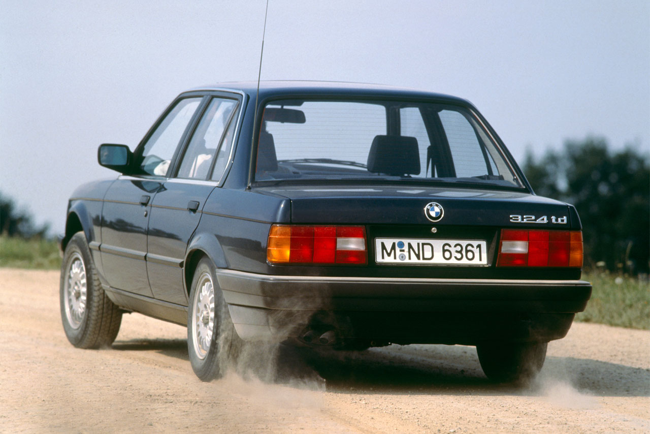 Dieselklassiker in Fahrt: Sicht auf Heck und Fahrerseite eines dunklen BMW E30 324td, der Staub auf einer unbefestigten Straße aufwirbelt.