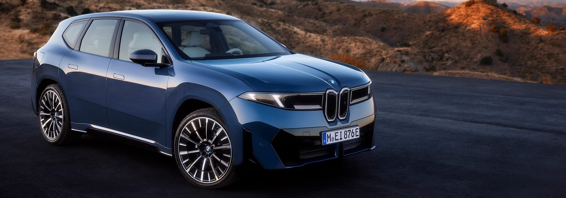 Der neue BMW iX3 in blau auf einer leeren Straße mit Sanddünen im Hintergrund.