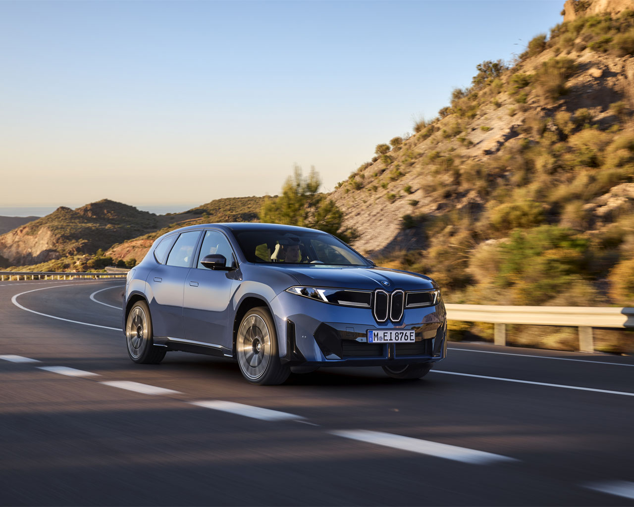 Ein BMW iX3 in blauer Farbe fährt auf einer kurvenreichen Straße, umgeben von Hügeln und Bäumen.