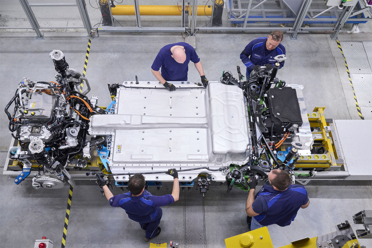 Blick von oben auf den Unterboden eines BMW iX1 im BMW Group Werk Regensburg: Vier Werker montieren Leitungen für die Motor-Getriebeeinheit.