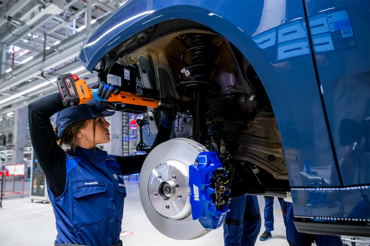 Eine Werkerin arbeitet in der Montage des BMW Group Werk in Debrecen mit einem Werkzeug vorne links am Fahrwerk eines BMW iX3.