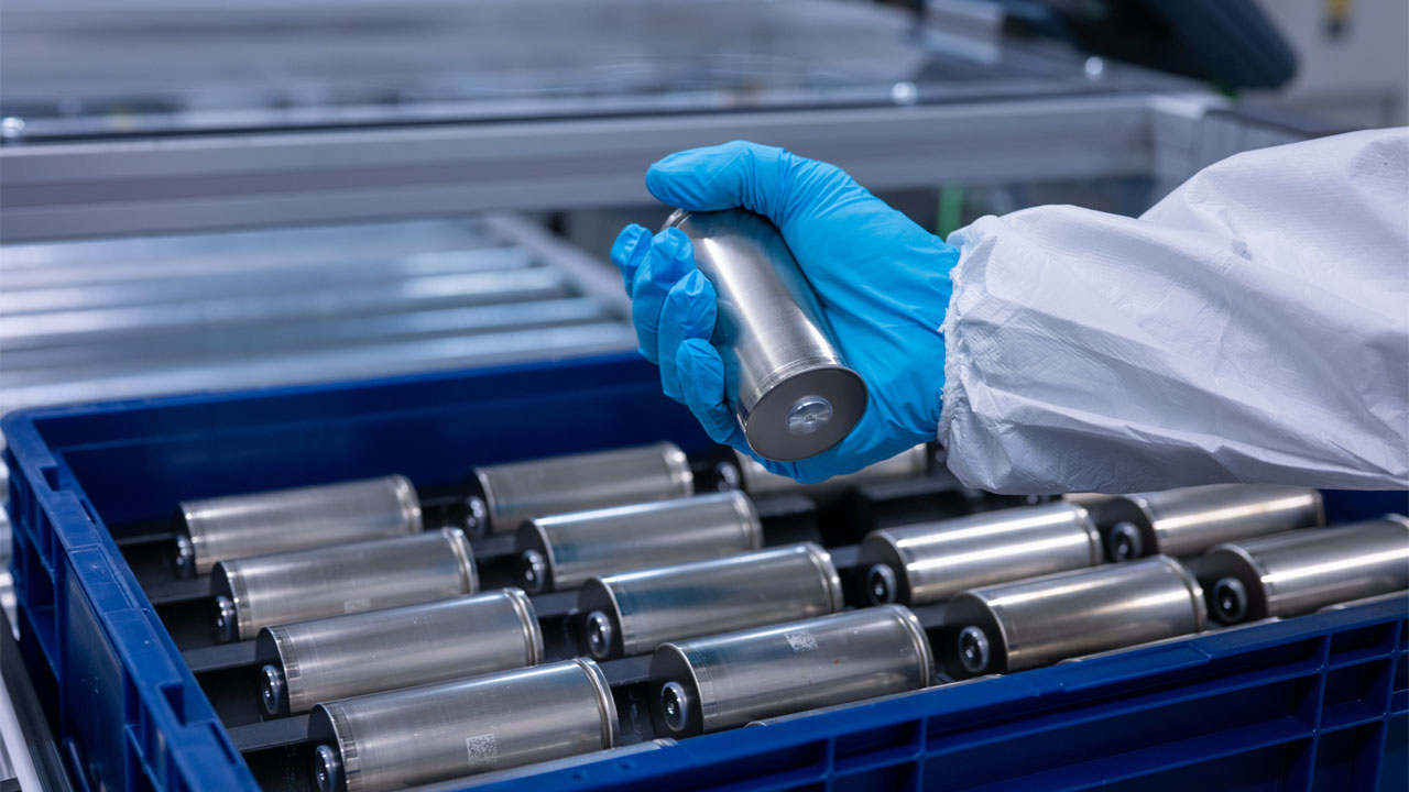 Fachkraft im Schutzanzug hält eine zylindrische Batteriezelle über eine blaue Transportkiste mit weiteren Zellen im BMW Group Kompetenzzentrum Batteriezellrecycling (CRCC).