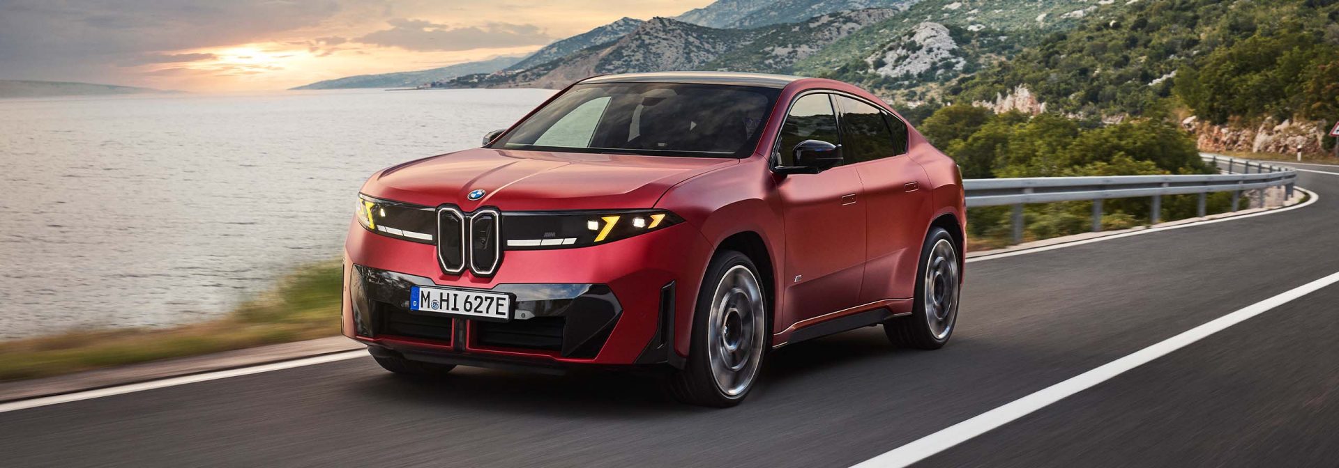 Schräg von vorn ist ein roter BMW iX4 M60 xDrive zu sehen, der auf einer Küstenstraße fährt. Im Hintergrund liegen das Meer und eine Bergkette im Abendlicht.