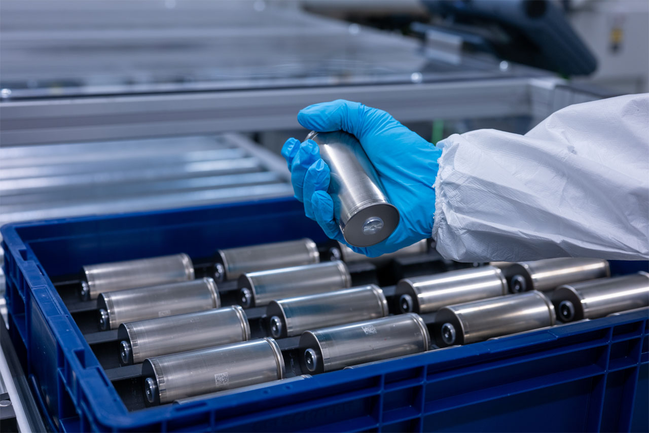 Fachkraft im Schutzanzug hält eine zylindrische Batteriezelle über eine blaue Transportkiste mit weiteren Zellen im BMW Group Kompetenzzentrum Batteriezellrecycling (CRCC).