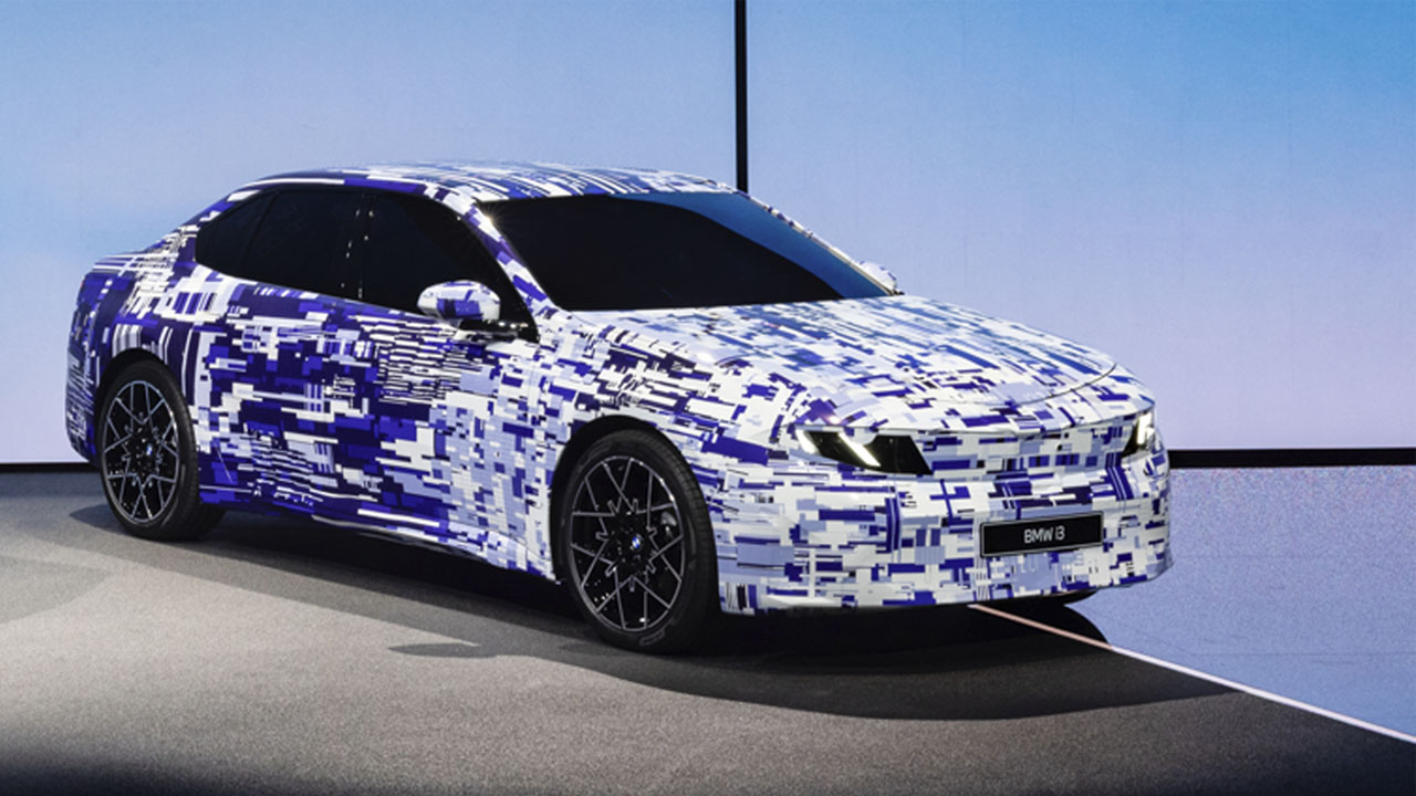 Vollelektrische Zukunft: Ein blau-weiß getarnter BMW i3 (Erlkönig) steht auf einer Präsentationsbühne.