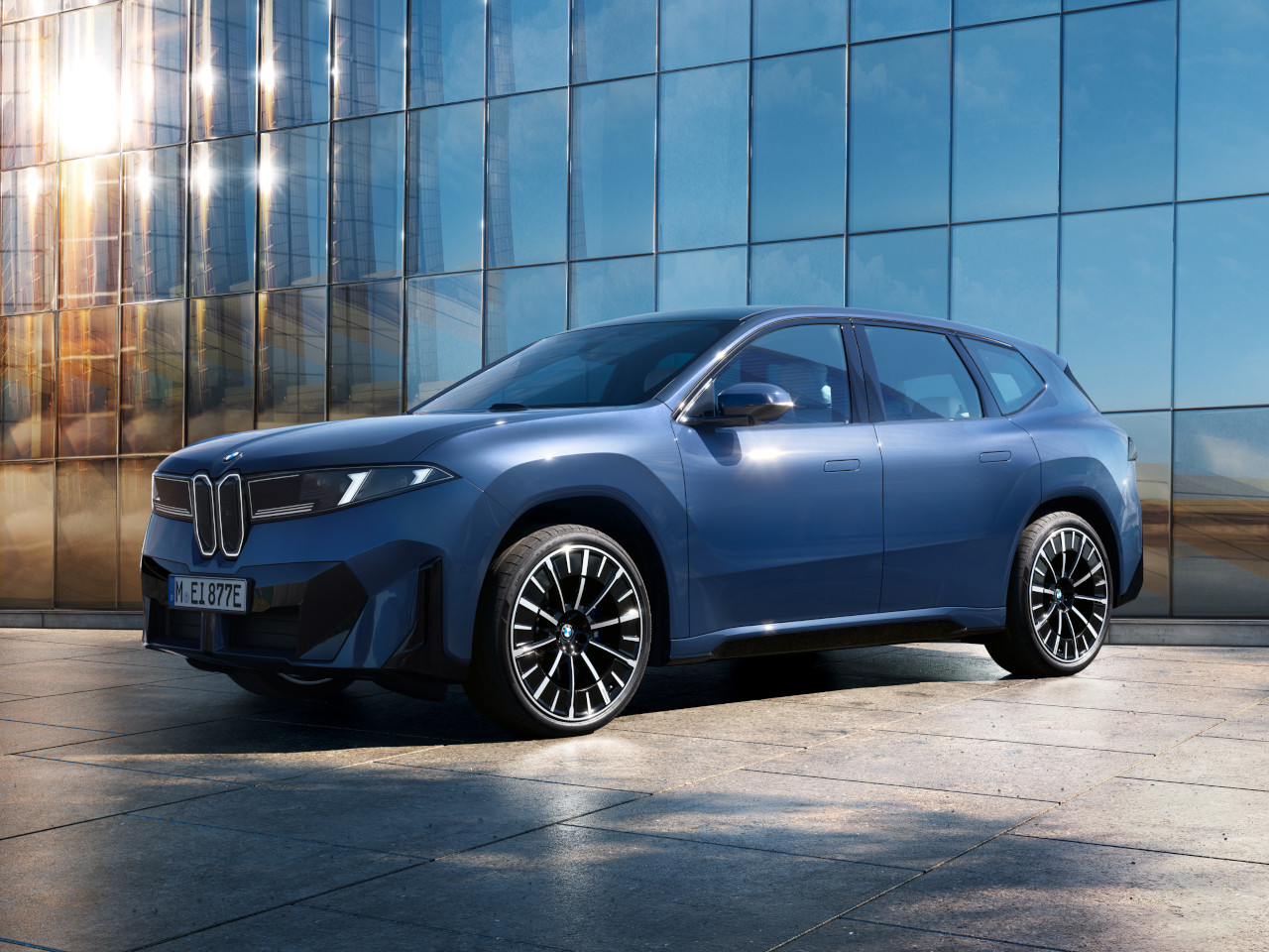 Seitenansicht des vollelektrischen BMW iX3, inszeniert vor moderner Glasfassade im Sonnenlicht.