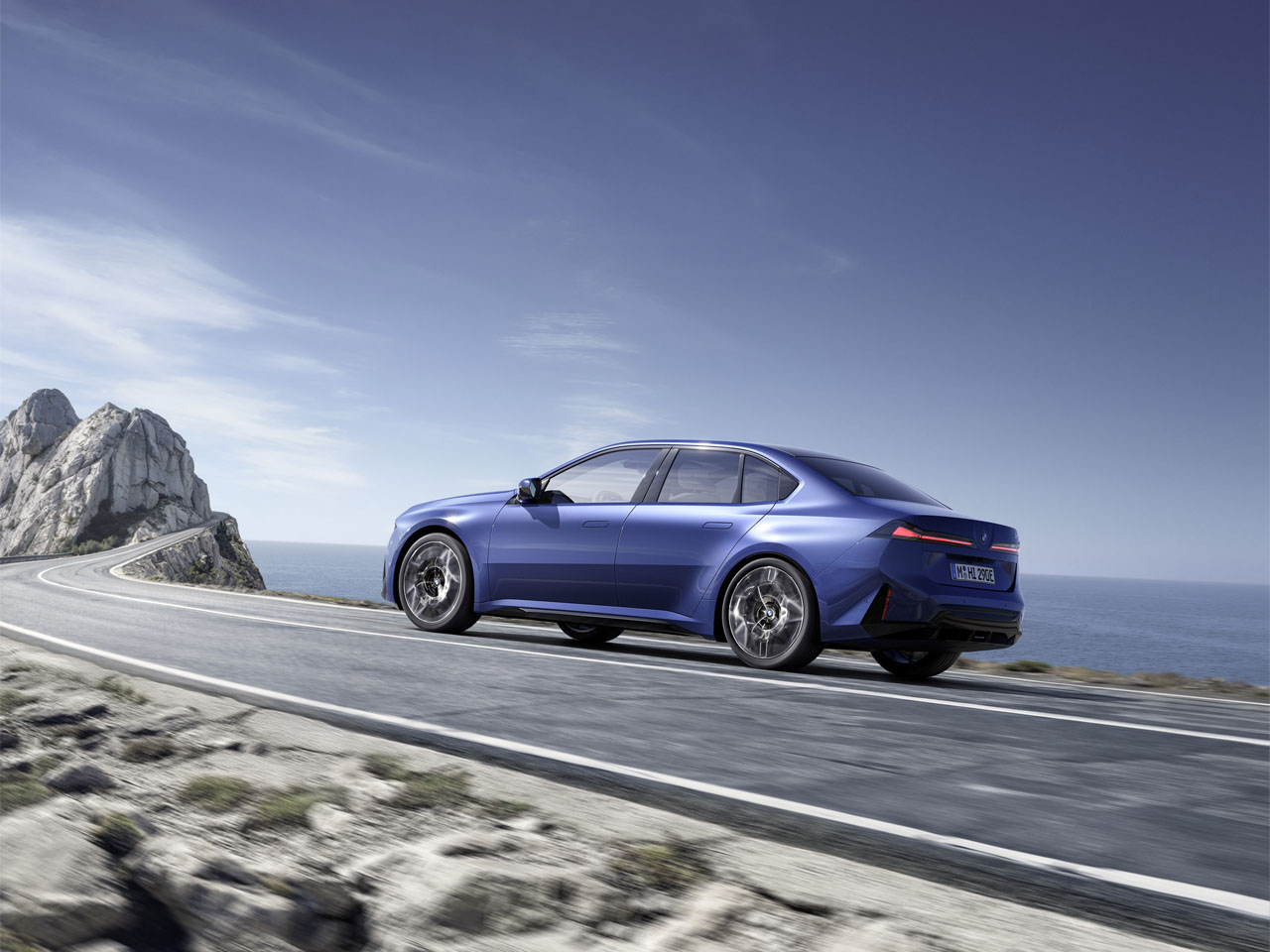 Blauer viertüriger Limousinenwagen fährt auf kurviger Küstenstraße mit Felsen und Meer im Hintergrund.