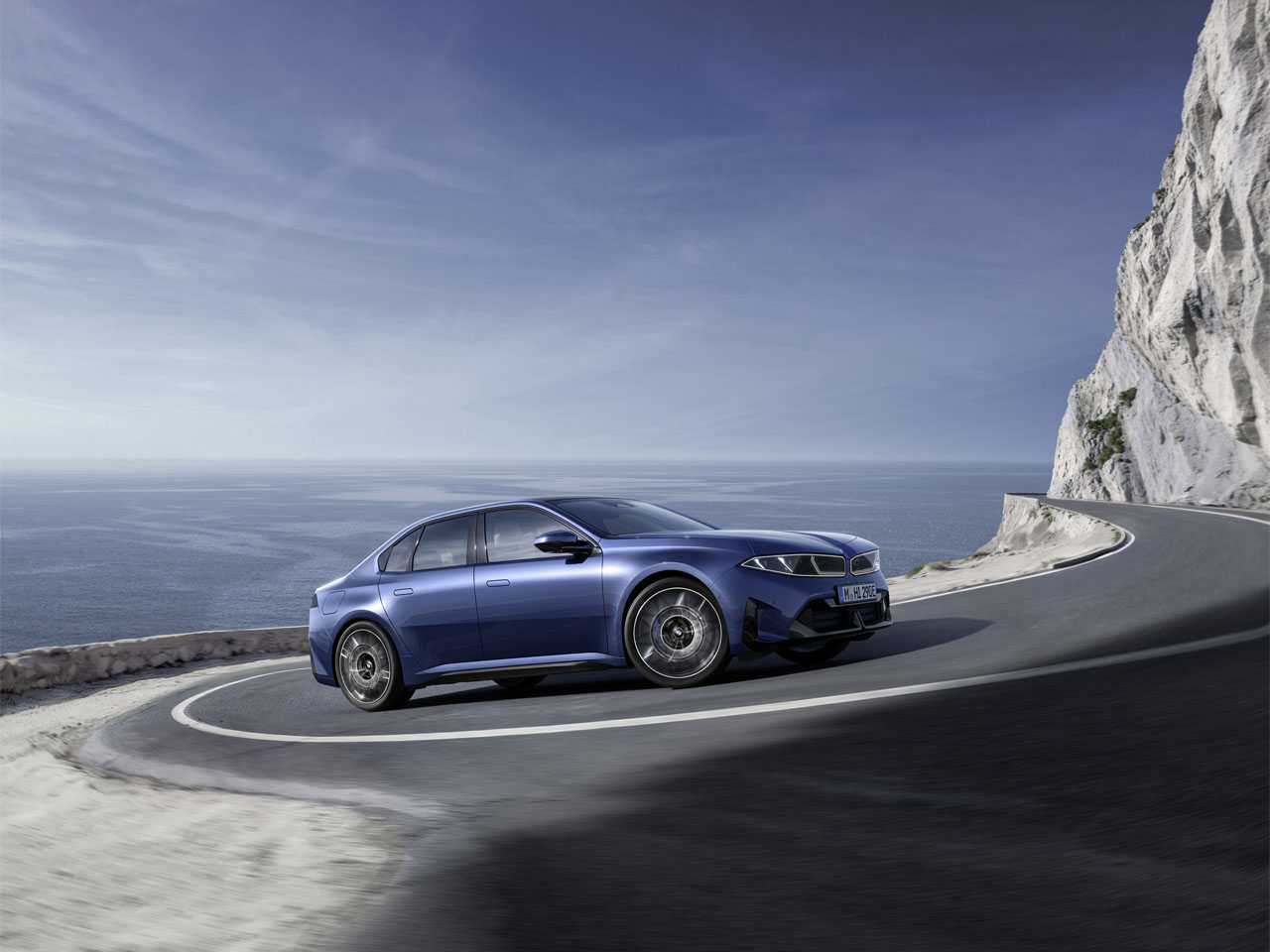 Blauer BMW der Neuen Klasse fährt auf einer kurvigen Küstenstraße neben einer Felswand.