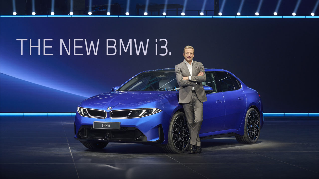 Ein Mann im Anzug lehnt mit verschränkten Armen an einem blauen BMW i3 vor einer Wand mit der Aufschrift 'THE NEW BMW i3'