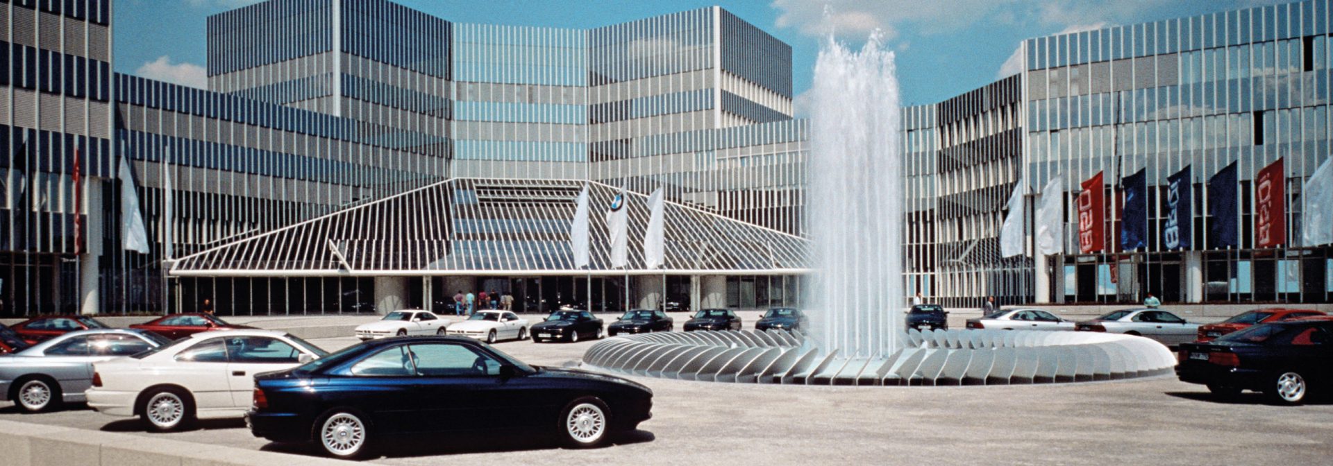Eingang zum BMW Group Forschungs- und Innovationszentrum mit geparkten Autos rund um den Springbrunnen.