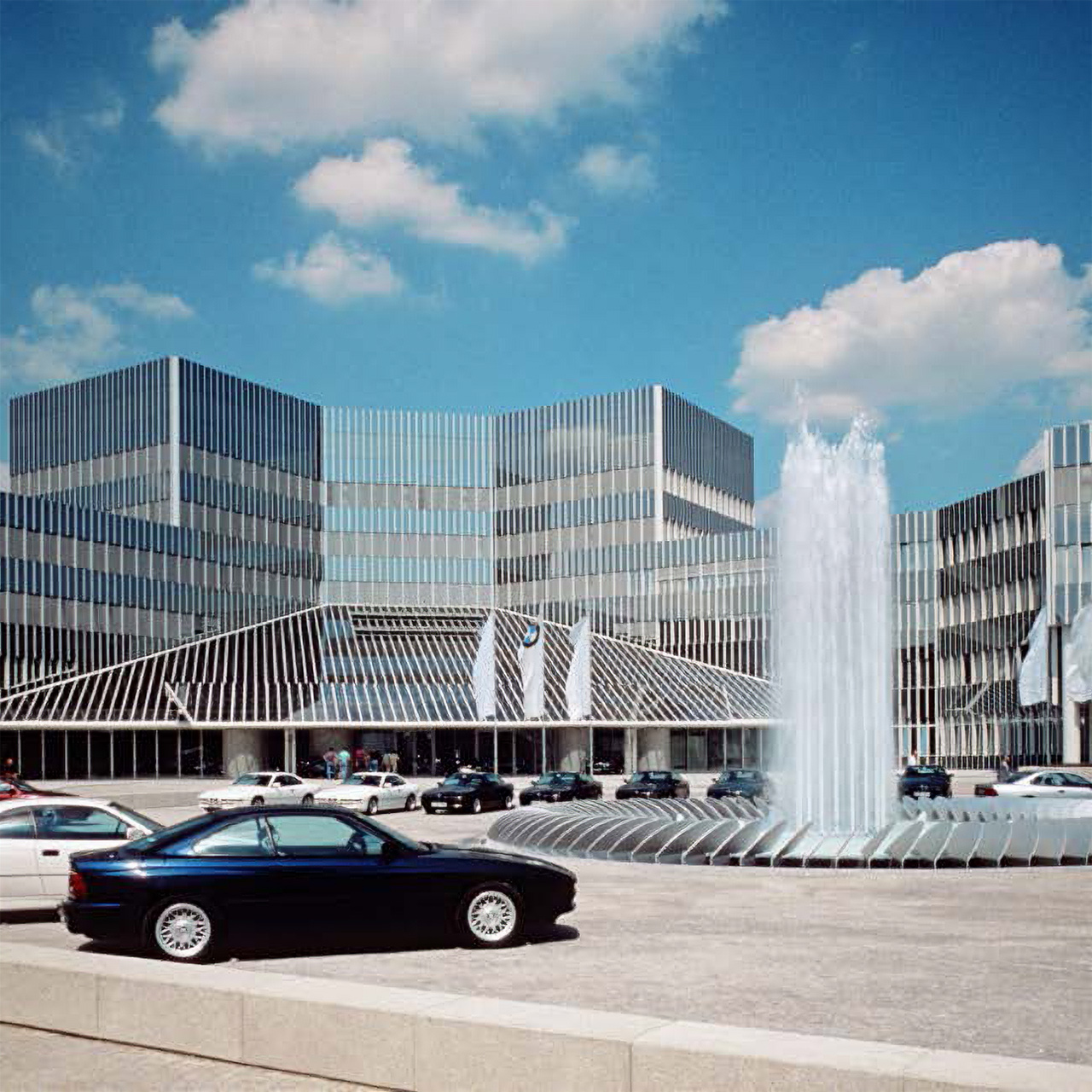 Eingang zum BMW Group Forschungs- und Innovationszentrum mit geparkten Autos rund um den Springbrunnen.