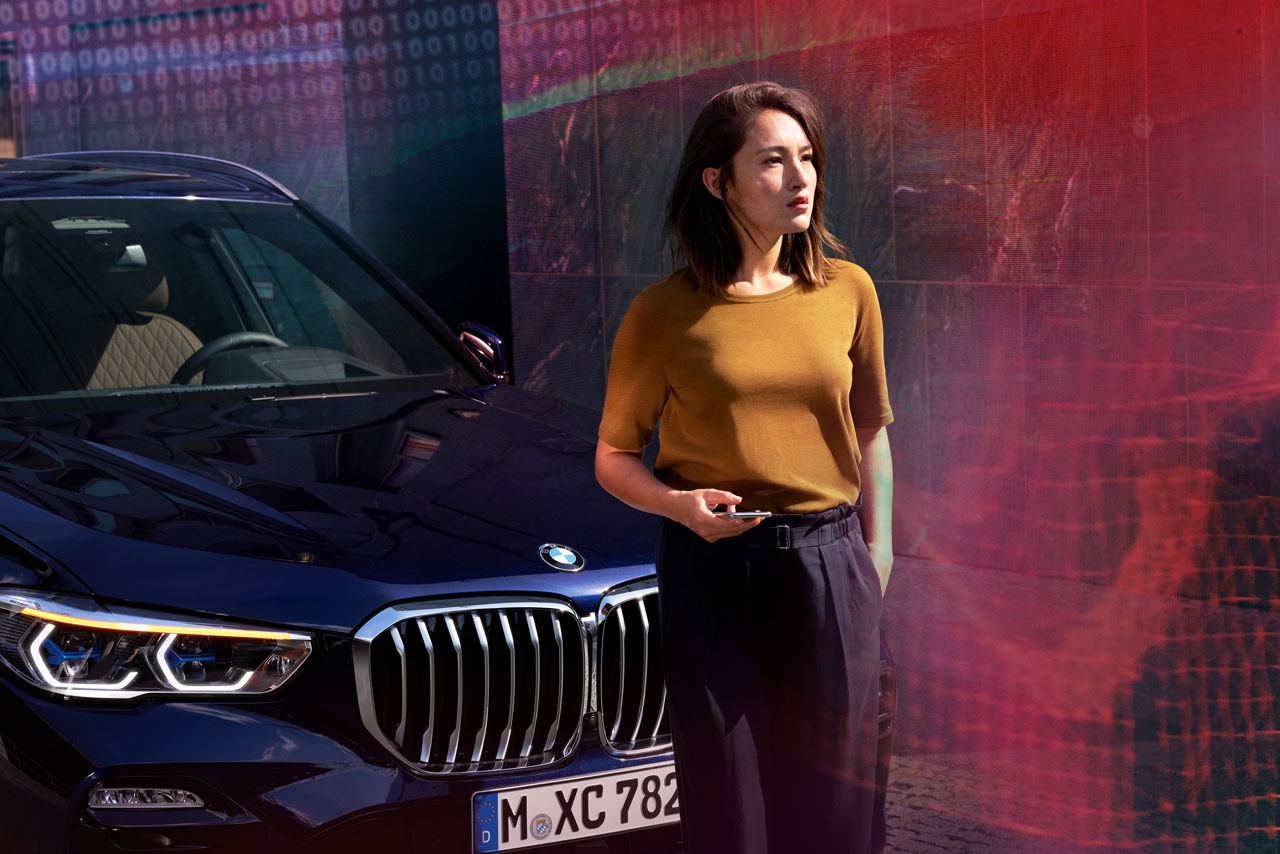 Eine Frau steht vor ihrem BMW und hält ein Smartphone in der Hand.