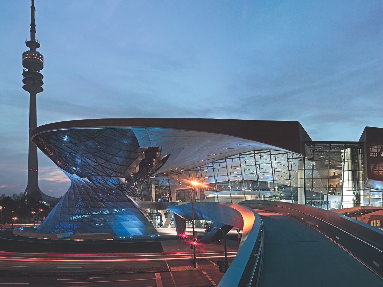 Evening view of BMW Welt with illuminated glass and metal structure, with Munich’s Olympic Tower in the background on the left.