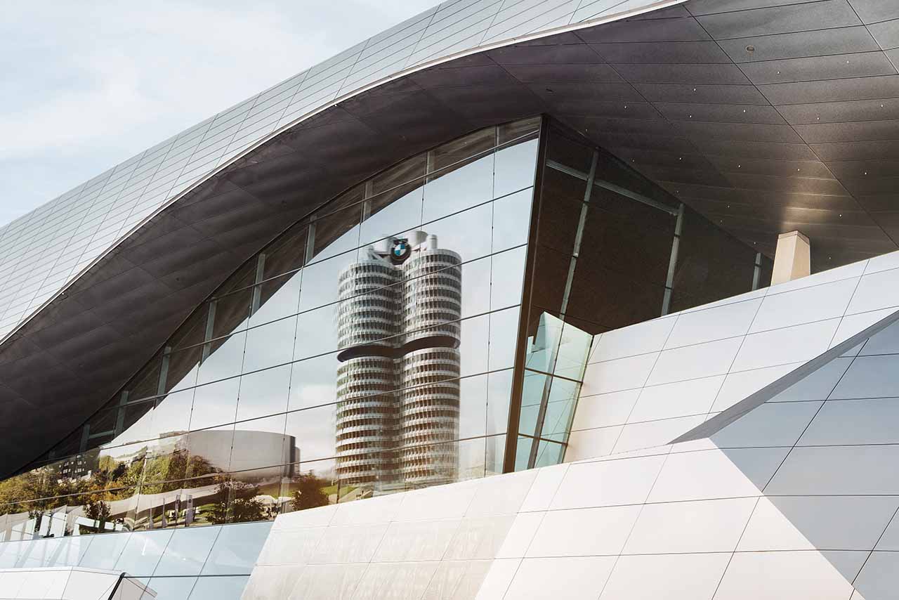 Reflection of a round high-rise building with BMW logo in a modern glass facade under a curved roof.