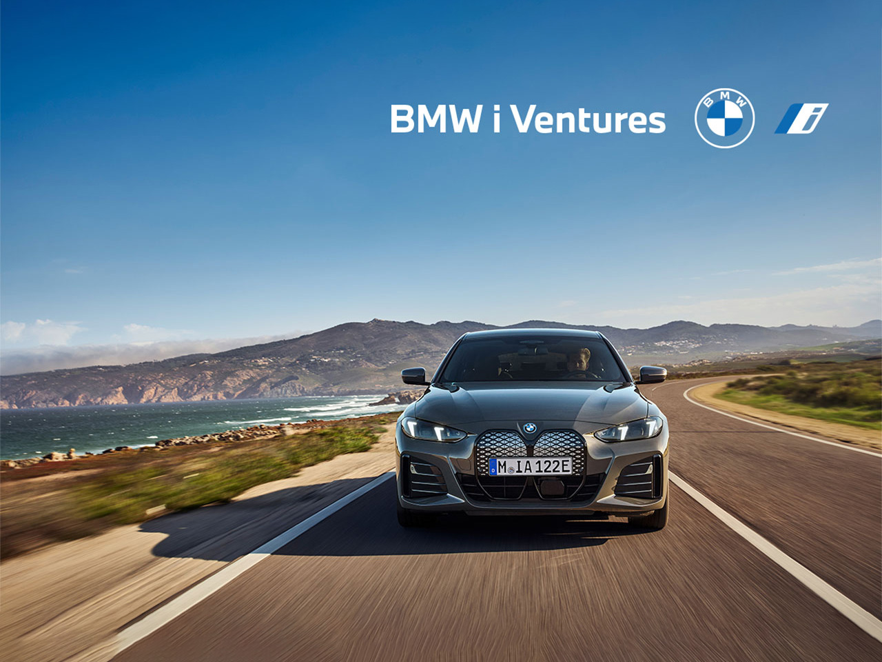 A gray BMW i4 drives on a winding coastal road. Mountains and the sea are visible in the background. Text: 'BMW i Ventures' and the BMW logo.