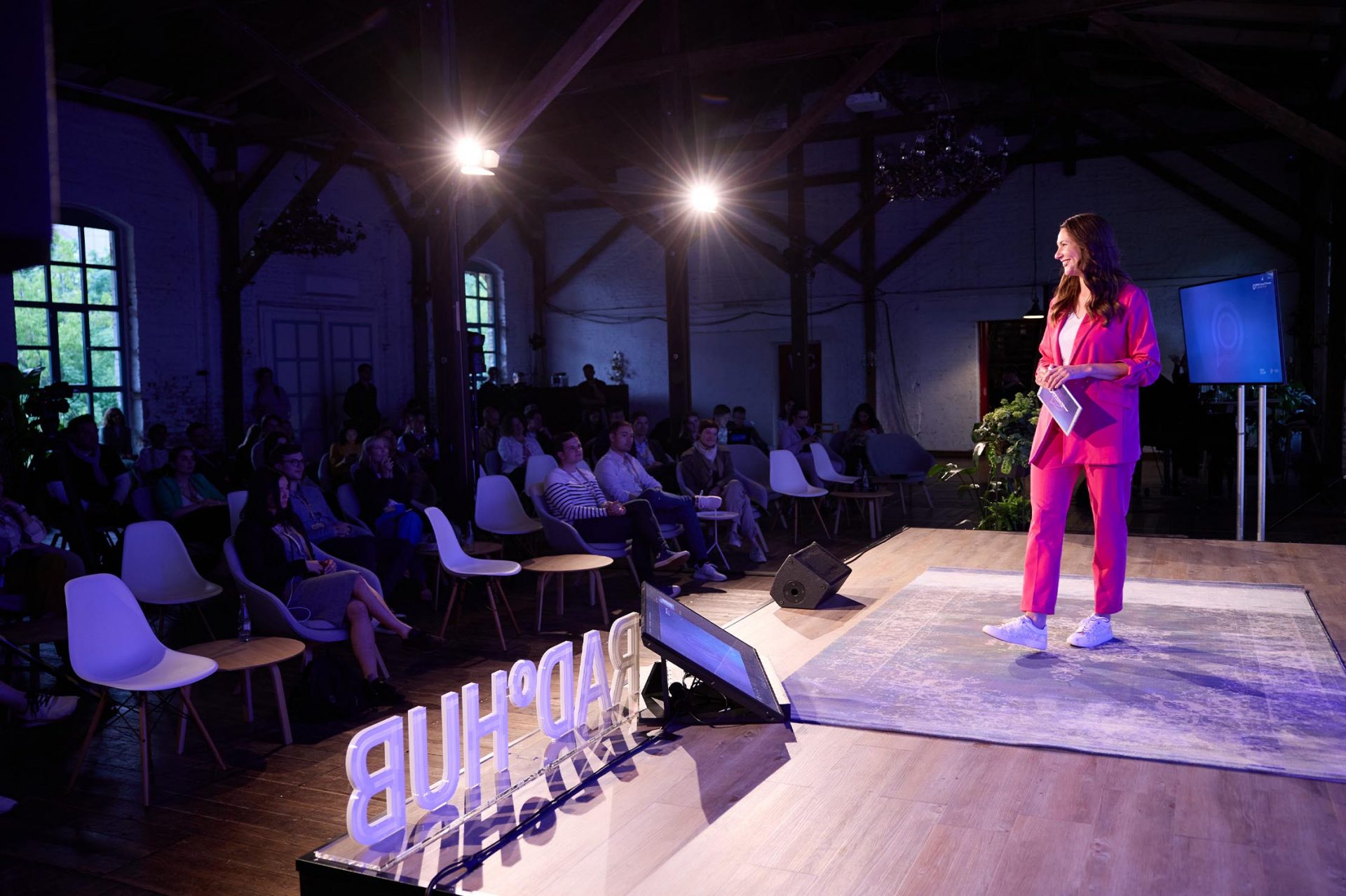   A speaker dressed in pink is addressing an audience on a conference stage. 