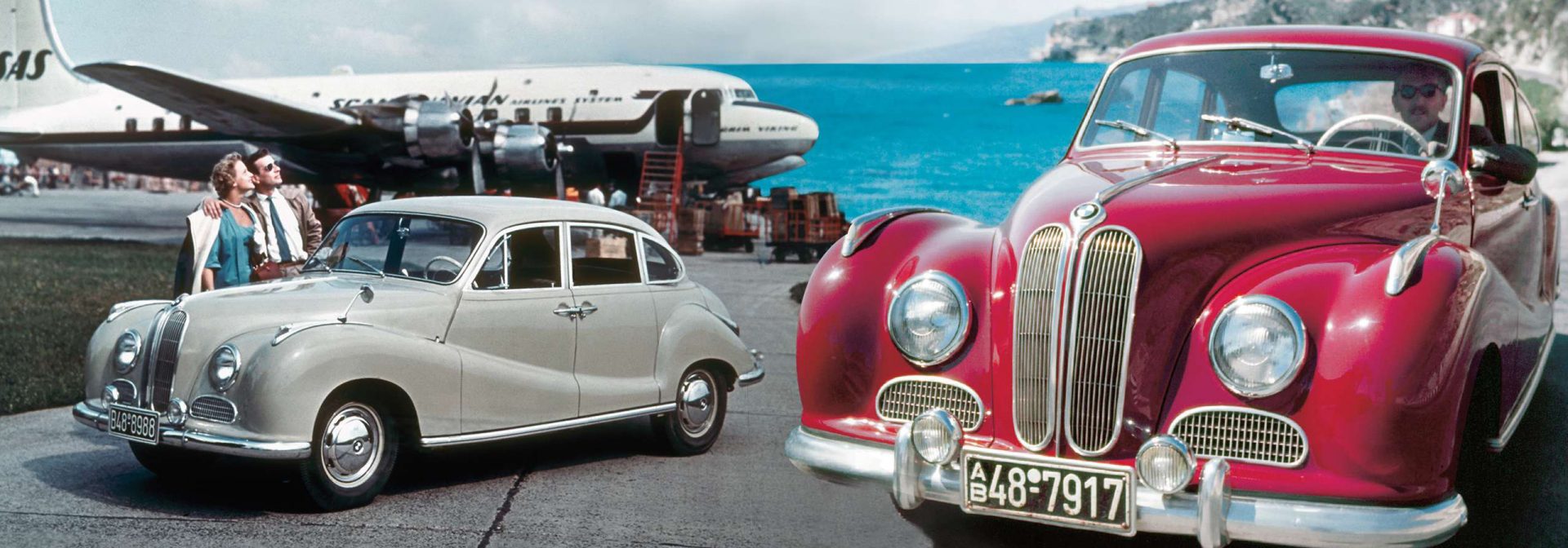 Two BMW 501, the first model produced after the war, one in grey and one in red. In the background there is a plane and the sea.