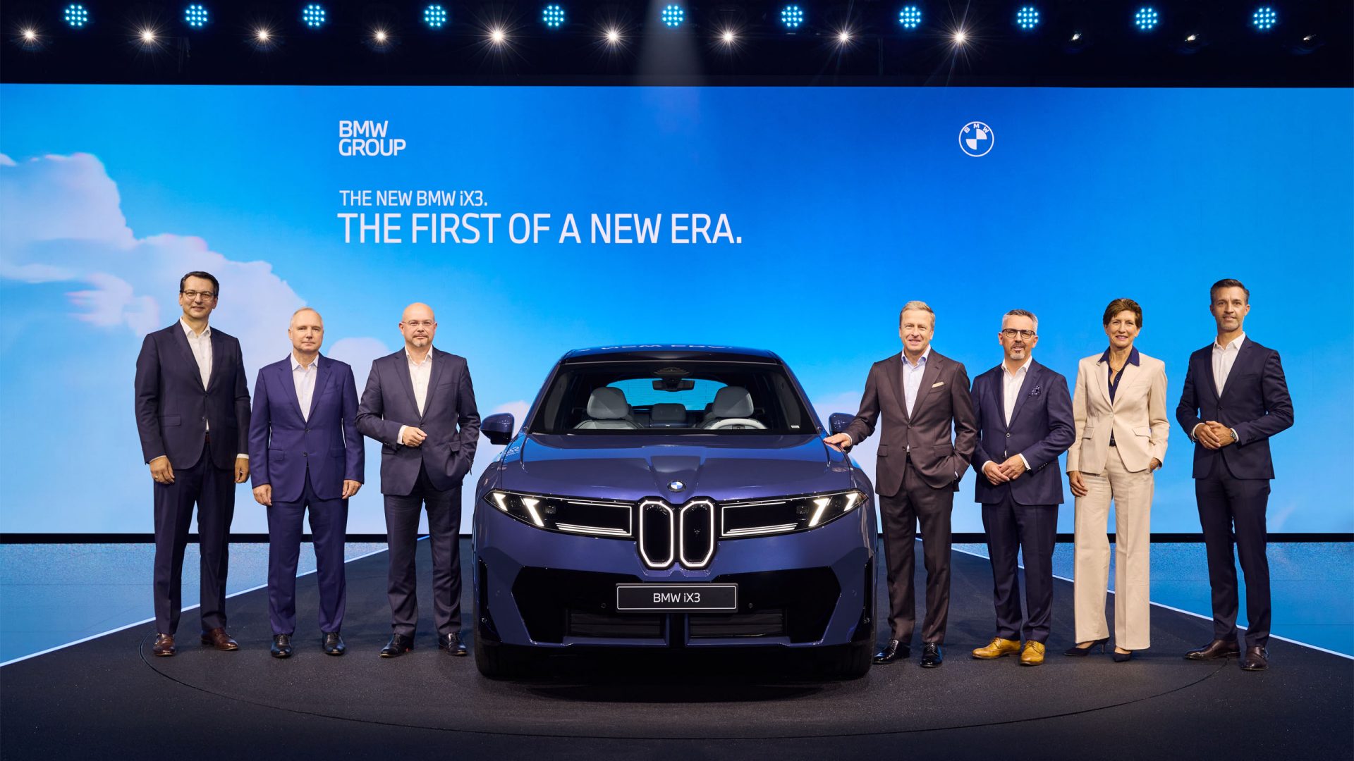 A group of six people stands beside a blue BMW iX3 on stage. The background displays the text 'THE FIRST OF A NEW ERA.'