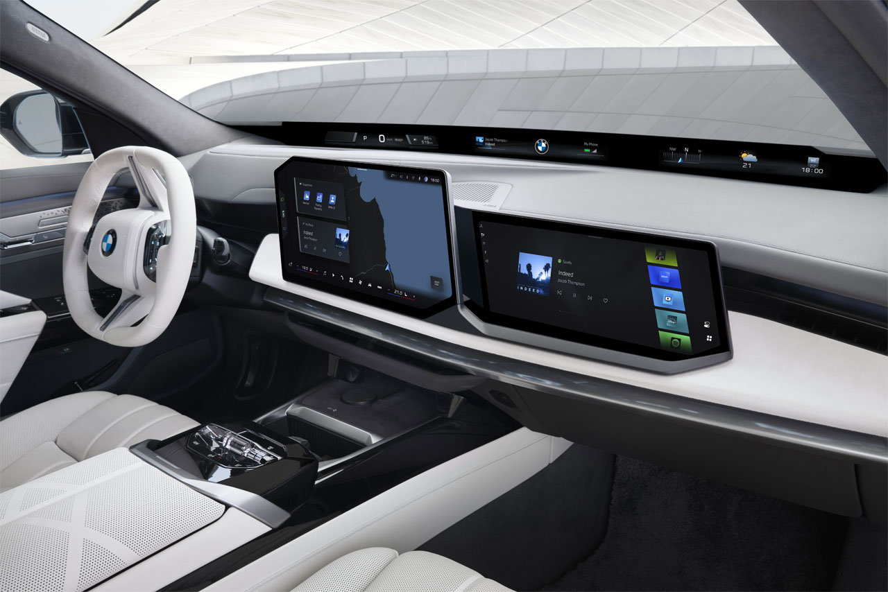 View across into the cockpit of a BMW 740 xDrive with a white steering wheel. The central display shows the navigation map, while the passenger-side display lists media offerings. The BMW Panoramic Vision presents driving information below the bottom edge of the windscreen.