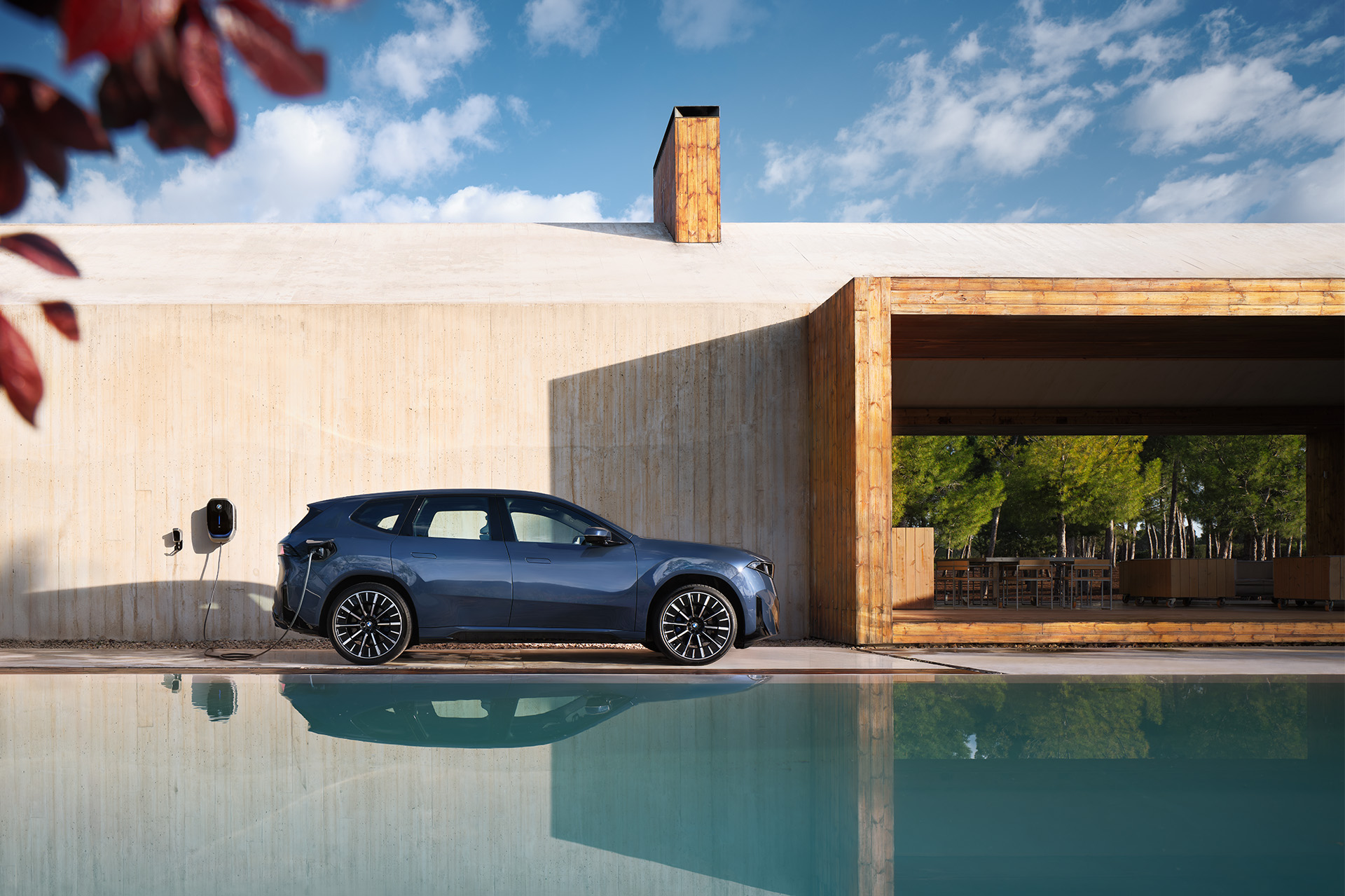 A blue BMW iX3 is being charged at a wall-mounted charging station of a futuristic-looking house.